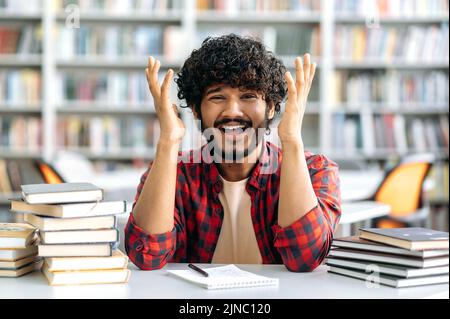 Un Indien perplexe ou un homme arabe, un étudiant de l'université, s'assoir à un bureau dans la bibliothèque entre les livres, regardant la caméra dans la déception de répandre ses bras sur les côtés, l'expression du visage émotionnel Banque D'Images