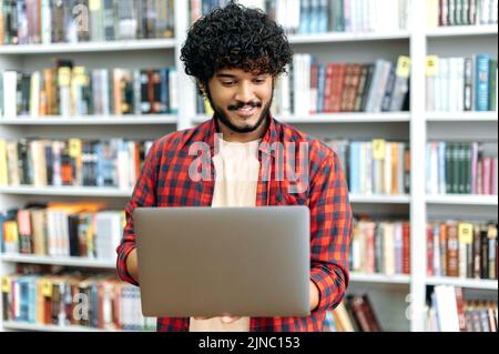 Elégant, moderne et aux cheveux mauresque, étudiant positif d'université arabe ou indienne, tenant un ordinateur portable ouvert dans ses mains, debout dans la bibliothèque, la navigation sur Internet, la recherche d'informations, souriant Banque D'Images