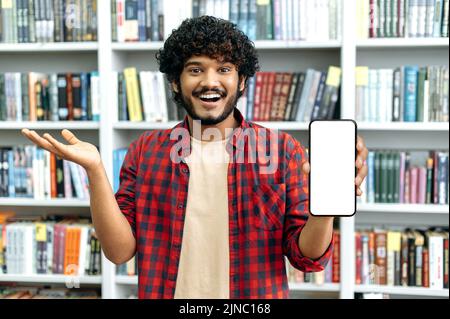 Un bon étudiant arabe ou indien, indépendant, montre un smartphone avec un écran blanc maquette, se tient dans la bibliothèque sur le fond des étagères, regarde l'appareil photo, sourit Banque D'Images