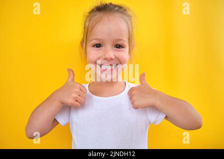 Bonne joyeuse petite fille d'âge préscolaire souriante montrant les pouces vers le haut fond jaune Banque D'Images