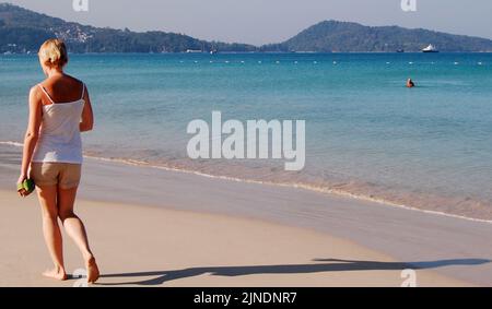 Une femme caucasienne marche le long d'une plage de sable tranquille en Thaïlande, en Asie, en début de matinée, jetant une longue ombre le long du sable. Un homme solitaire se tient dans la mer. Banque D'Images