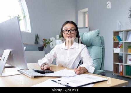 Travailler avec des documents. Portrait d'une jeune belle femme d'affaires asiatique comptable travaille avec des documents et des rapports. Assis au bureau, écrivant.il regarde la caméra, sourit Banque D'Images