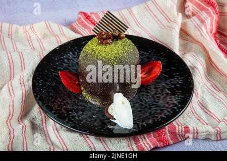 Un gâteau sucré. Gâteau au chocolat glacé, une pièce, sur une assiette noire avec gâteau à la pistache. Il est servi sur une assiette de crème glacée et de fraises. Banque D'Images