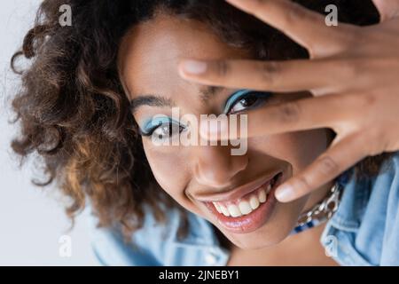 femme afro-américaine gaie avec maquillage obscurcissant visage avec la main isolée sur bleu Banque D'Images