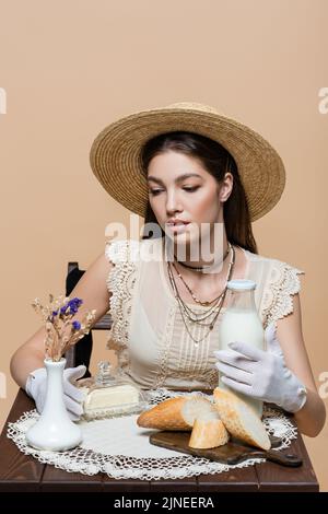 Modèle tendance en chapeau de soleil tenant le lait et le beurre près du pain sur la table isolée sur beige Banque D'Images