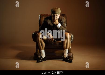 Portrait cinématographique d'un jeune homme, gangster anglais rétro de style 1920s portant un costume et une casquette isolés sur un arrière-plan vintage sombre. Affaires, famille, art Banque D'Images