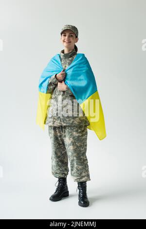 Soldat heureux couvrant avec drapeau ukrainien sur fond gris, image de stock Banque D'Images