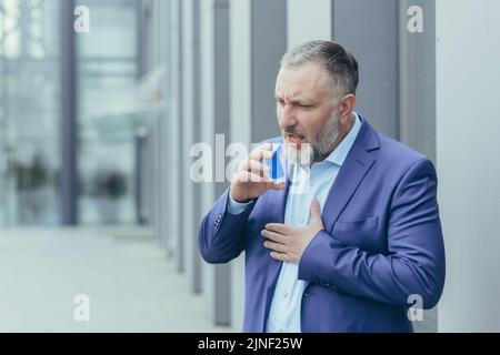 Un homme d'affaires âgé atteint de cheveux gris souffre d'une crise d'asthme difficile à respirer, un homme à l'extérieur du bâtiment de bureau dans les vêtements d'affaires utilise un inhalateur pour soulager sa respiration Banque D'Images