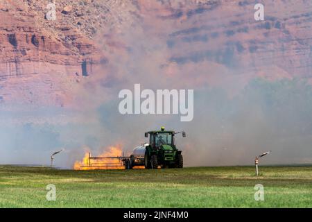 Un tracteur tirant un brûleur de propane brûle les mauvaises herbes dans un champ de foin après avoir coupé la luzerne dans un ranch de l'Utah. Banque D'Images