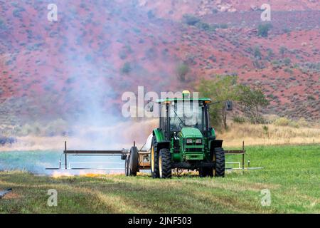 Un tracteur tirant un brûleur de propane brûle les mauvaises herbes dans un champ de foin après avoir coupé la luzerne dans un ranch de l'Utah. Banque D'Images