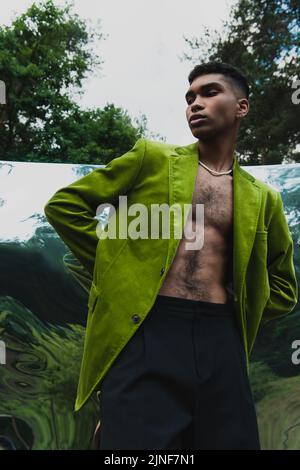 homme afro-américain tendance dans un blazer vert debout avec les mains derrière le dos près de la surface du miroir dans la forêt, image de stock Banque D'Images