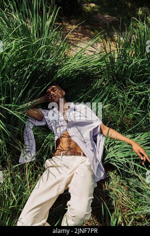 vue en grand angle de l'homme afro-américain élégant avec les yeux fermés se détendant sur l'herbe verte, image de stock Banque D'Images