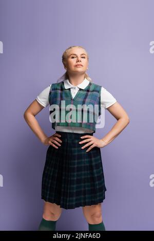 Étudiant confiant en uniforme à carreaux tenant les mains sur les hanches isolées sur violet, image de stock Banque D'Images