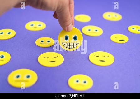 Gros plan de la main tenant un smiley jaune visage emoji entre les émoticônes tristes sur fond violet Banque D'Images