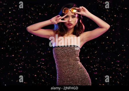 Belle femme asiatique portant une robe lurex et des lunettes de soleil posant pour la photographie tout en dansant en boîte de nuit, confetti colorés volant dans l'air Banque D'Images