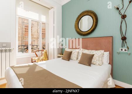 Chambre avec lit double, couvre-lits bruns assortis avec coussins, têtes de lit capitonnées en bois, miroir circulaire décoratif et balcon avec vue Banque D'Images