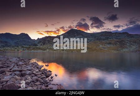 Vue sur le coucher du soleil avec une lumière orange spectaculaire derrière les sommets des montagnes Retezat et reflétée dans le lac Bucura. Photo prise le 6th août 2022 le Banque D'Images