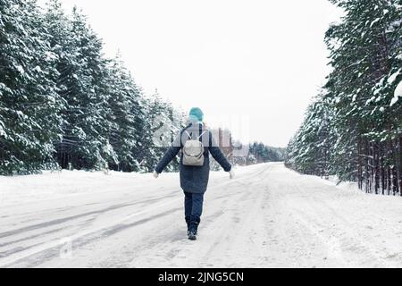 une fille en manteau gris et un bonnet tricoté bleu, avec un sac à dos gris sur son dos, marche le long d'une route d'hiver Banque D'Images