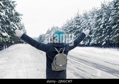 fille dans un manteau gris, avec un sac à dos gris derrière son dos, se tient sur le côté de la route, dans une belle forêt d'hiver Banque D'Images