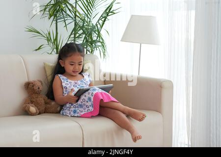 Petite fille assise sur un canapé avec une tablette numérique sur les genoux Banque D'Images