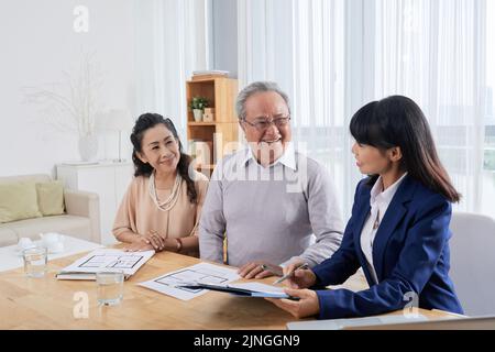 Sympathique agent immobilier asiatique assis à un bureau spacieux avec des clients âgés et les aidant à remplir le formulaire pour louer un appartement Banque D'Images