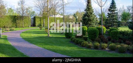 Aménagement paysager dans le parc avec chemin en pierre et sapins, clôture verte avec thuya, buissons et arbres. Paysage de printemps panorama. Banque D'Images