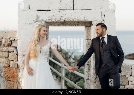 Portrait d'un couple de mariage souriant debout près de l'arche ancienne blanche. Jeune femme mariée tenant la main avec le marié en costume. Banque D'Images