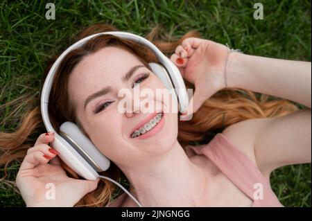 Une jeune femme aux cheveux rouges se trouve sur l'herbe et écoute de la musique. Banque D'Images