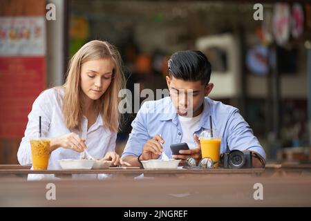 Jeune couple de touristes mangeant de la nourriture de rue asiatique, homme utilisant son smartphone Banque D'Images