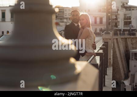 Vue latérale d'un couple de mariage attrayant se tenant sur la rambarde en acier du pont au coucher du soleil. Jeune mariée embrassant avec marié. Banque D'Images