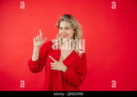 Portrait d'une jeune femme en robe rouge vif debout et pointant les doigts vers le haut isolés sur fond rouge. Publicité moderne, copie gratuite Banque D'Images