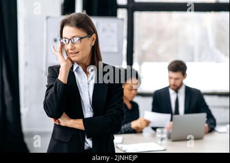 Portrait d'élégante femme d'affaires de race blanche avec des lunettes, en costume formel, chef de l'entreprise, chef d'entreprise, debout dans un bureau moderne, regardant à côté, souriant Banque D'Images