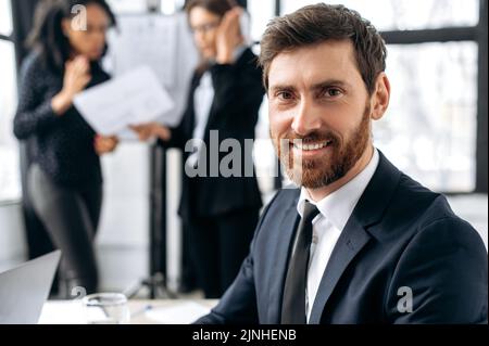 Homme d'affaires élégant et confiant, pdg de la société, assis au bureau pendant le brainstorming d'entreprise, regarde la caméra, sourit. Portrait d'un dirigeant d'affaires caucasien attrayant en costume lors d'une réunion d'information Banque D'Images