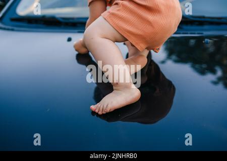 Un portrait en gros plan des pieds d'un bébé qui essaie de se déplacer se trouve sur le capot d'une voiture. Bonne famille, enfance. Voyage en voiture. Bonne famille à l'extérieur. Banque D'Images