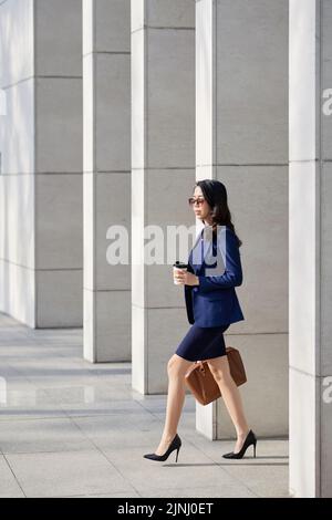 Vue de profil d'un jeune Manager confiant en tenue de formal tenant une tasse de papier de café parfumé en main tout en quittant le bâtiment de bureau après une dure journée de travail Banque D'Images