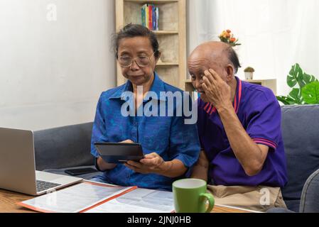 Les personnes âgées asiatiques s'assoient et tiennent une calculatrice pour les frais d'assurance ou médicaux et un ordinateur sur le bureau. Banque D'Images