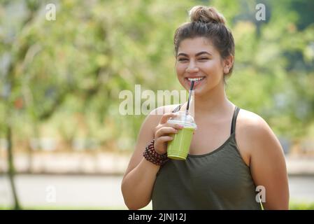 Jeune femme souriante portant un superbe appareil photo tout en buvant un smoothie de paille et en profitant d'une vue pittoresque sur un parc public verdoyant Banque D'Images