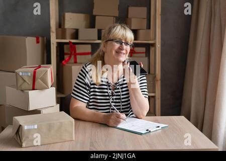 Femme mature propriétaire de petite entreprise utilisant le téléphone mobile vérifiant boîte colis. Un vendeur d'entrepreneur plus âgé tenant un téléphone cellulaire dicte l'assistant vocal Banque D'Images