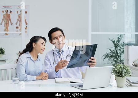 Beau chirurgien d'âge moyen assis au bureau et montrant une image radiologique au patient senior tout en discutant d'un traitement possible, à l'intérieur d'un bureau moderne en arrière-plan Banque D'Images