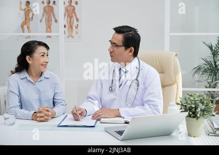 Patient âgé souriant assis à un bureau à côté d'un médecin d'âge moyen et répondant aux questions pendant qu'il remplit son dossier médical, à l'intérieur d'un bureau moderne en arrière-plan Banque D'Images