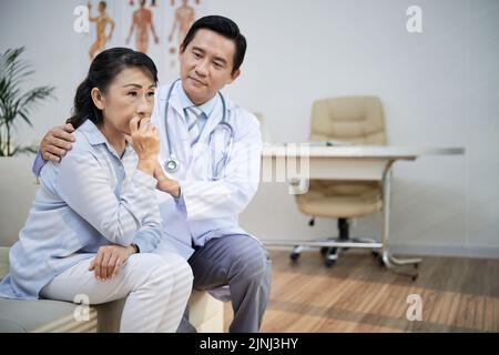 Beau médecin asiatique assis sur un canapé de bureau moderne et apaisant la triste femme âgée après l'annonce du diagnostic Banque D'Images
