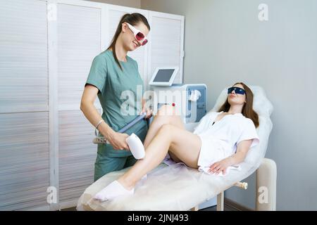 Jeune femme couché sur une table médicale dans une clinique de beauté ayant l'enlèvement de cheveux au laser dans un salon de beauté professionnel.Esthéticienne féminine en uniforme à l'aide d'une machine d'épilation laser Banque D'Images