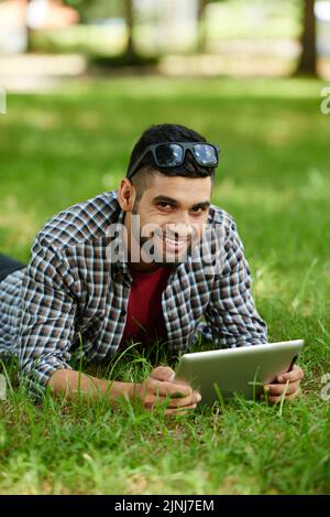 Homme indien souriant allongé sur une pelouse verte et posant pour la photographie tout en surfant sur Internet sur une tablette numérique moderne, portrait tourné Banque D'Images