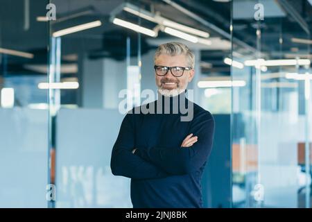 Portrait d'un bon patron à cheveux gris en lunettes, homme d'affaires travaillant dans un bureau moderne debout près d'une fenêtre souriant et regardant un appareil photo avec des bras croisés, investisseur satisfait en lunettes Banque D'Images