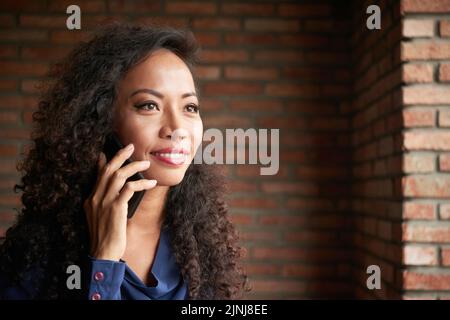 Portrait de la tête et des épaules d'une femme asiatique attrayante qui regarde loin avec un grand sourire tout en parlant à un ami sur un smartphone, mur de briques sur l'arrière-plan Banque D'Images