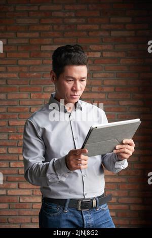 Portrait d'un entrepreneur asiatique confiant utilisant une tablette numérique tout en travaillant sur un projet prometteur, mur de brique de bureau moderne sur fond Banque D'Images