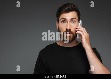 un homme choqué dans un t-shirt noir parlant sur un smartphone isolé sur une image grise Banque D'Images