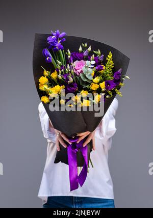 Bouquet de roses jaunes et eustoma pourpre dans un emballage en papier noir dans les mains d'une femme debout sur fond gris. Une femme inconnue tient la flo Banque D'Images