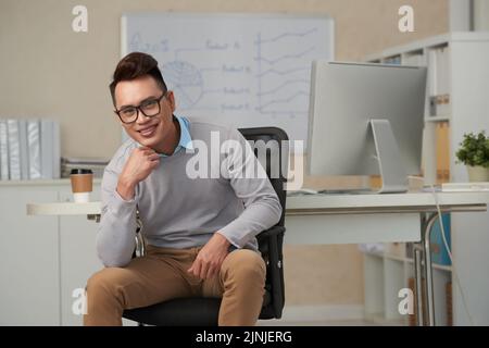 Beau jeune entrepreneur souriant assis sur un fauteuil dans son bureau Banque D'Images