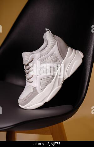 Affichage des détails sur les baskets. Gros plan d'une sneaker blanche debout sur le dos d'une chaise noire en studio sur fond beige. Photo promotionnelle. S Banque D'Images